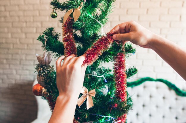 Manos decorando el árbol de Navidad con oropel rojo Primer plano de un pino verde decorado con adornos brillantes y lazos Concepto de felicidad de invierno de vacaciones