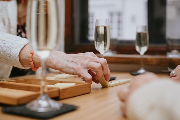 Manos con dedos delgados y pulsera naranja posiblemente de anciana con chaqueta blanca jugando dominó en la mesa con vasos llenos de vino o champán