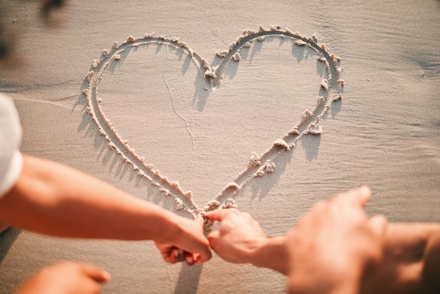 Manos dibujando y corazón en la arena de la playa para el cuidado y la pareja con amabilidad o apoyo junto con emoji Dibuja el amor o escribe el símbolo en forma de amoroso cuidado o línea en el romance