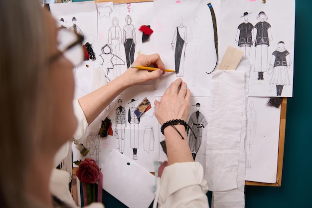 Manos de diseñador de moda sosteniendo lápiz y esbozando ropa en papel clavado en la pared con dibujos y esquises adjuntos