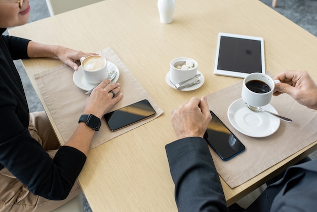 Manos de dos corredores elegantes discutiendo noticias financieras o proyectos de su empresa comercial por taza de café mientras están sentados a la mesa en el restaurante