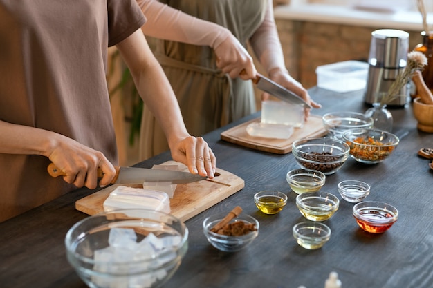 Foto manos de dos mujeres jóvenes cortando masa de jabón duro en cubos sobre tablas de madera antes de hacer productos cosméticos naturales en casa