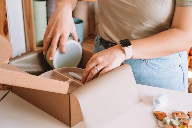 Foto manos de empresaria recortadas empaquetando y envolviendo el envío con platos de silicona para niños en caja de cartón tienda de taller