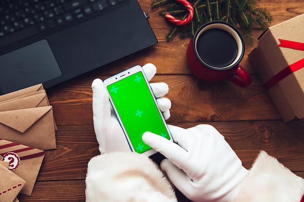 Manos enguantadas de santa claus sosteniendo un teléfono. Chromakey. Junto a él hay una computadora portátil, una taza, un regalo y ramas de abeto. Vista superior.