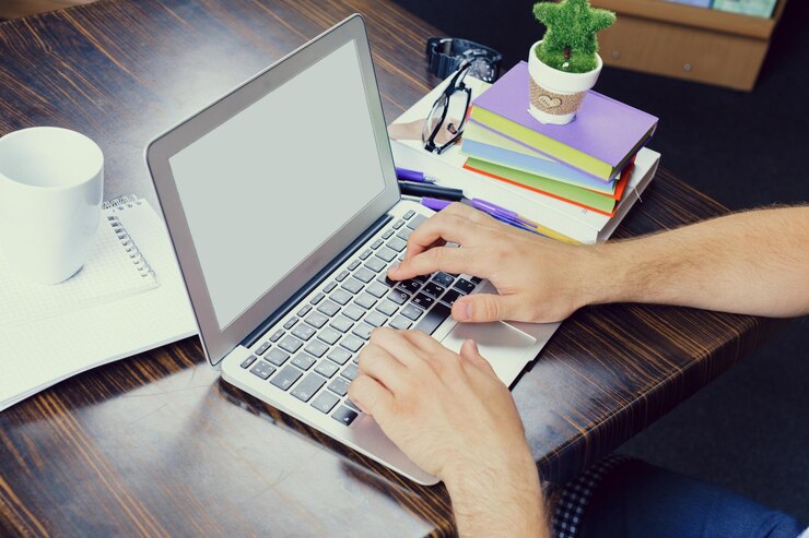 Foto las manos escribiendo en una computadora portátil en un escritorio de madera con un vaso, gafas y cuadernos de colores cerca en un espacio de trabajo acogedor
