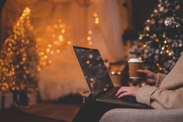 Manos escribiendo en una computadora portátil con un hermoso árbol de Navidad brillante en el fondo creando una atmósfera acogedora y festiva en el interior