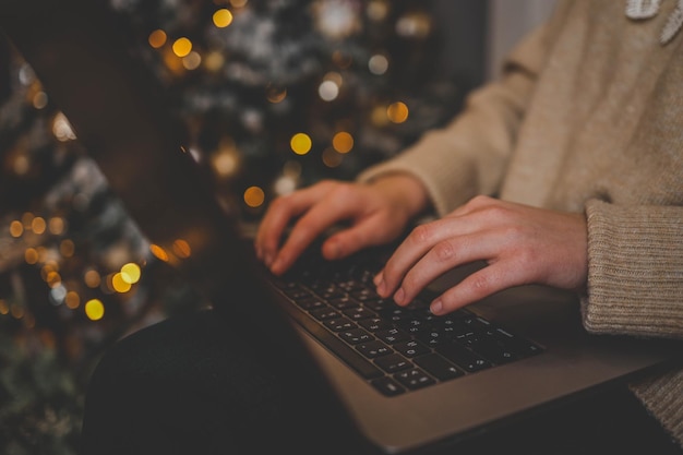 Manos escribiendo en una computadora portátil con un hermoso árbol de Navidad brillante en el fondo creando una atmósfera acogedora y festiva en el interior