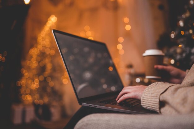 Manos escribiendo en una computadora portátil con un hermoso árbol de Navidad brillante en el fondo creando una atmósfera acogedora y festiva en el interior