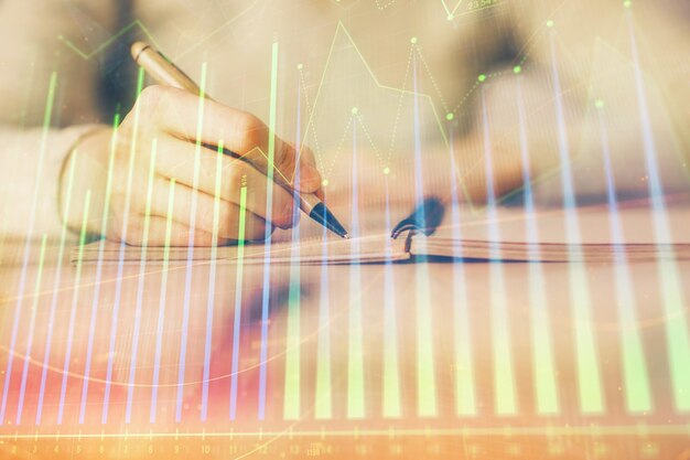 Manos escribiendo información sobre el mercado de valores en el bloc de notas gráficos de divisas hologramas en el frente concepto de investigación exposición múltiple