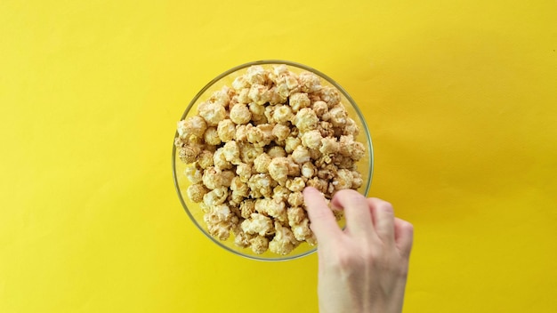 Las manos de la familia agarran palomitas de maíz. Recipiente redondo de vidrio con palomitas de maíz dulce sobre fondo amarillo. vista superior.