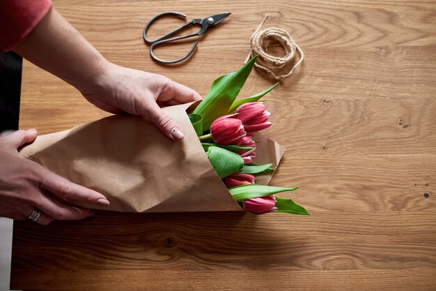 Manos femeninas arreglando el ramo de tulipanes rosados en la mesa de madera, lugar de trabajo de pasatiempo florístico, negocios, bricolaje, concepto de regalo de primavera, desde arriba.