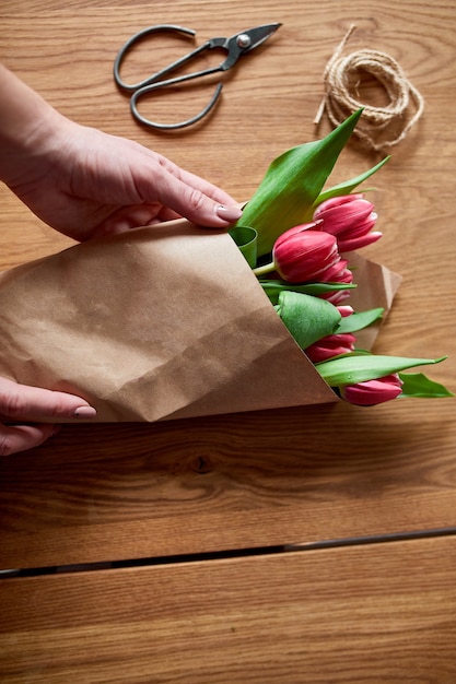 Manos femeninas arreglando el ramo de tulipanes rosados en la mesa de madera, lugar de trabajo de pasatiempo florístico, negocios, bricolaje, concepto de regalo de primavera, desde arriba.