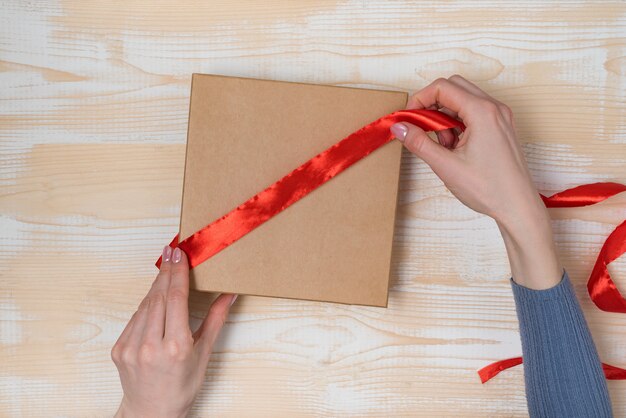 Las manos femeninas atan una cinta roja en una caja de regalo, mesa de madera, vista superior