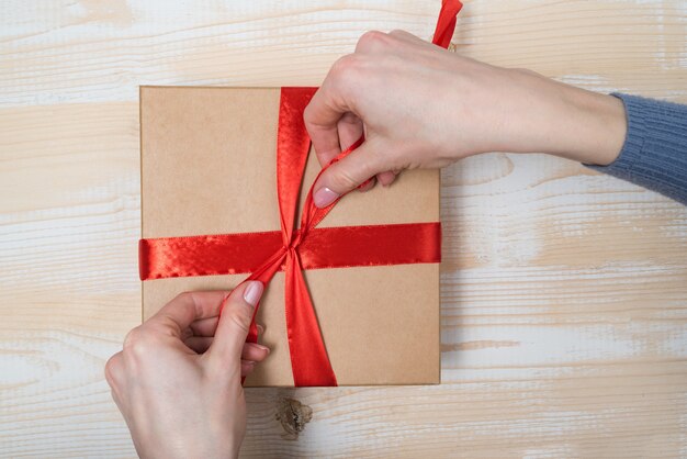 Las manos femeninas atan un lazo de cinta roja en una caja de regalo. Mesa de madera. Vista superior