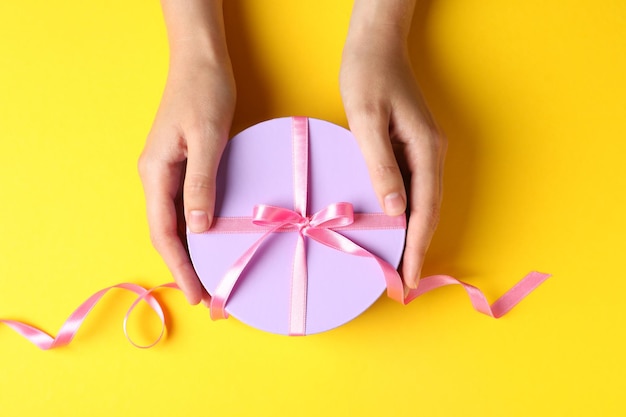 Manos femeninas y caja de regalo redonda con lazo sobre fondo amarillo