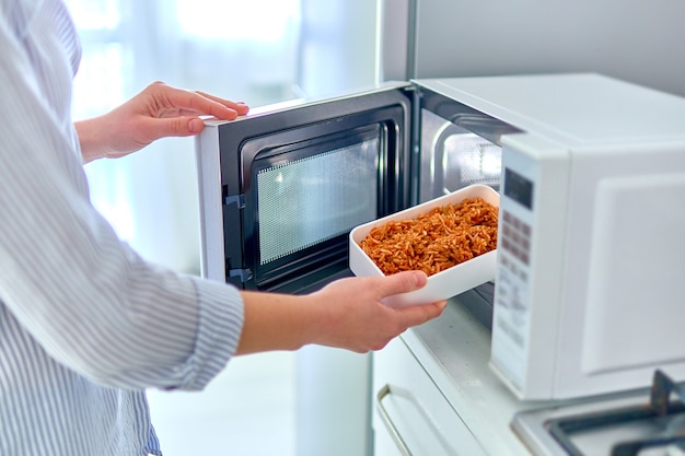 Manos femeninas calentando un recipiente de comida en el moderno horno de microondas para el almuerzo en casa