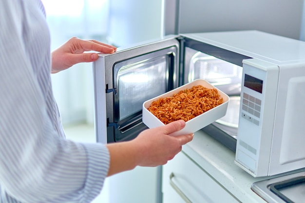 Manos femeninas calentando un recipiente de comida en el moderno horno de microondas para el almuerzo en casa