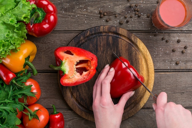 Manos femeninas cortando pimienta en la mesa, vista superior. Sobre la mesa hojas de lechuga, pimiento, un vaso de jugo de tomate, una tabla de madera y un cuchillo