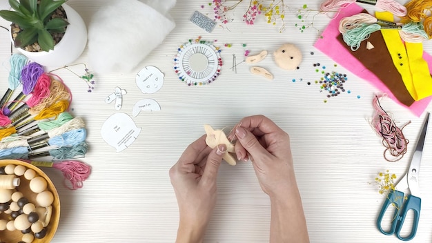 Foto manos femeninas cosen una muñeca de fieltro hecha a mano sobre fondo de mesa de madera blanca