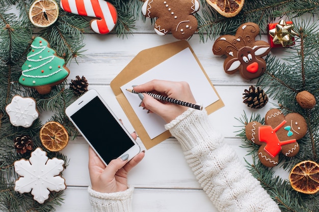 Las manos femeninas escriben una tarjeta de Navidad acogedora en un fondo de gengerbread y de los regalos.