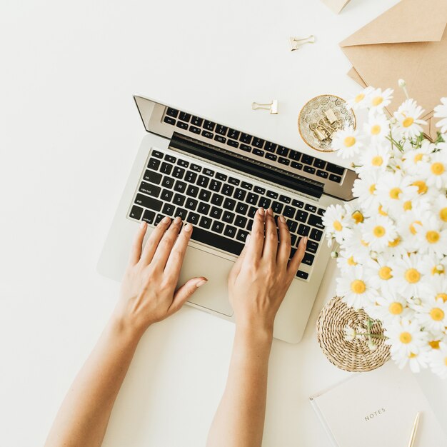 Manos femeninas escribiendo composición. Espacio de trabajo de escritorio de oficina en casa con computadora portátil, ramo de flores de manzanilla y margarita en blanco