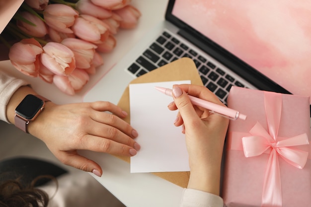 Manos femeninas escribiendo deseos en papel para San Valentín o cumpleaños con caja de regalo y portátil en el escritorio, vista superior.