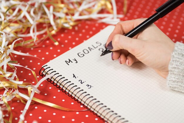 Manos femeninas escribiendo Mis objetivos 2020 en un cuaderno Fondo rojo y oropel Concepto de Año Nuevo