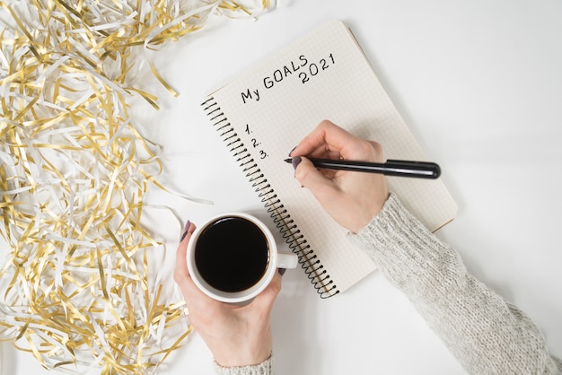 Manos femeninas escribiendo Mis objetivos 2021 en un cuaderno. Taza de café sobre la mesa, vista superior.