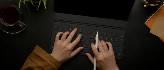 Manos femeninas escribiendo en el teclado de la tableta en el oscuro escritorio de oficina con agenda y decoración