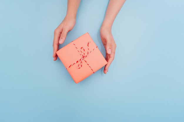 Las manos femeninas están sosteniendo una caja de regalo rosa con una cinta delgada como regalo para Navidad, Año Nuevo, Día de la Madre o aniversario en un fondo de mesa azul, vista superior Lugar para el texto.