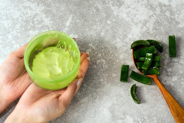 Manos femeninas con gel de aloe y una cuchara con rodajas de hojas de aloe sobre un fondo gris Fabricación de cosméticos naturales en casa