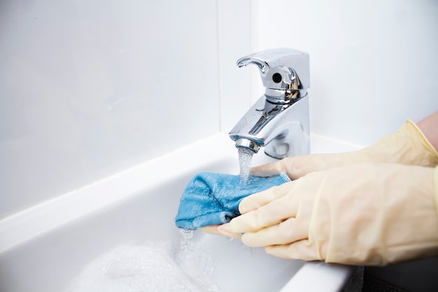 Manos femeninas en guantes bajo un chorro de agua con un trapo. Trapo azul en el agua. Grifo, agua, manos, trapo. Limpieza en húmedo.