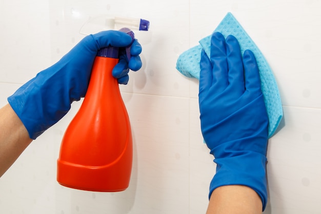 Manos femeninas en guantes de goma azul sostiene una botella de detergente y una servilleta de microfibra. La mujer está lavando la pared con azulejos blancos en el baño. Herramientas y equipo de limpieza