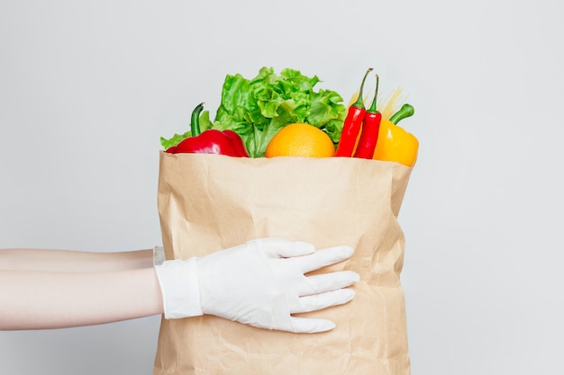 Las manos femeninas en guantes médicos sostienen una bolsa de papel con alimentos, verduras, pimienta, chile, hierbas frescas aisladas, entrega segura de alimentos