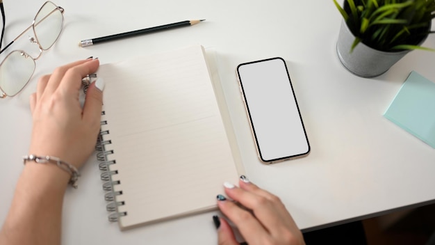 Manos femeninas con maqueta de páginas de cuaderno vacías y maqueta de pantalla en blanco de teléfono inteligente en mesa blanca