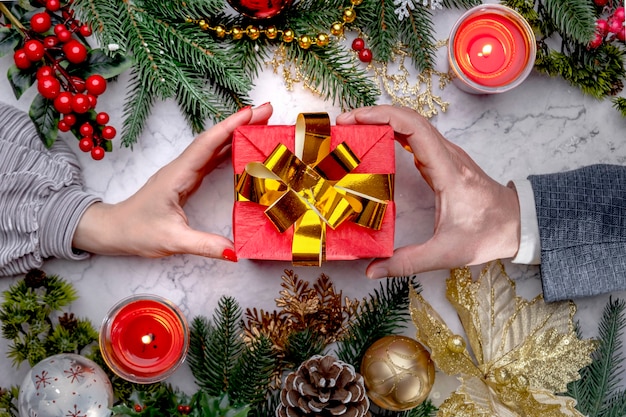 Manos femeninas y masculinas dando caja de regalo roja de Navidad con un lazo dorado sobre mármol blanco. Vista superior plana Lay. Ramas de abeto de año nuevo, conos, bolas, adornos de oro de lujo. Ánimo de vacaciones