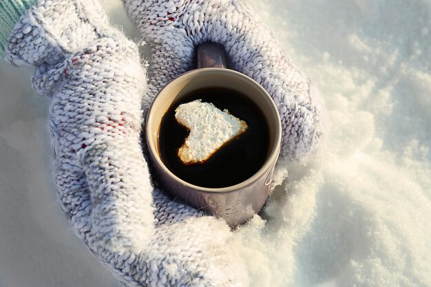 Manos femeninas en mitones calientes sosteniendo una taza de café caliente con malvavisco de corazón sobre fondo nevado