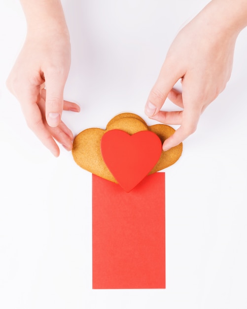 Las manos femeninas ponen las galletas de la forma del corazón con la tarjeta de felicitación roja en un fondo blanco. Símbolo de amor acogedor y fondo del día de San Valentín, simulacro