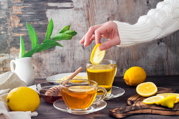 Las manos femeninas ponen una rodaja de limón en una taza de té, limones y un tazón de miel sobre una mesa de madera. Vitamina Nutrición Orgánica