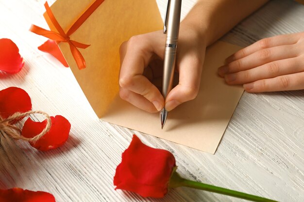 Manos femeninas que firman una tarjeta de regalo para el Día de San Valentín en un fondo blanco de madera