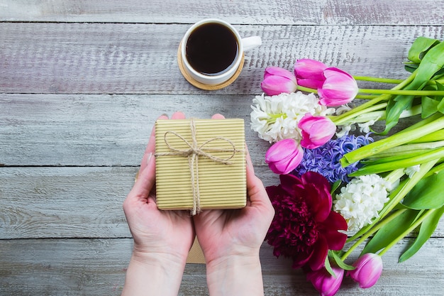 Manos femeninas que sostienen un regalo o una caja actual, flores tulipanes, peonía, jacinto y taza de café negro