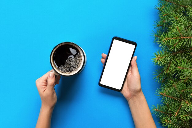 Manos femeninas que sostienen el teléfono móvil negro con la pantalla blanca en blanco y la taza de café.