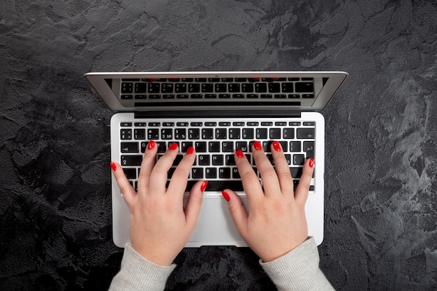 Foto manos femeninas con uñas rojas escribiendo en la computadora portátil sobre fondo negro.