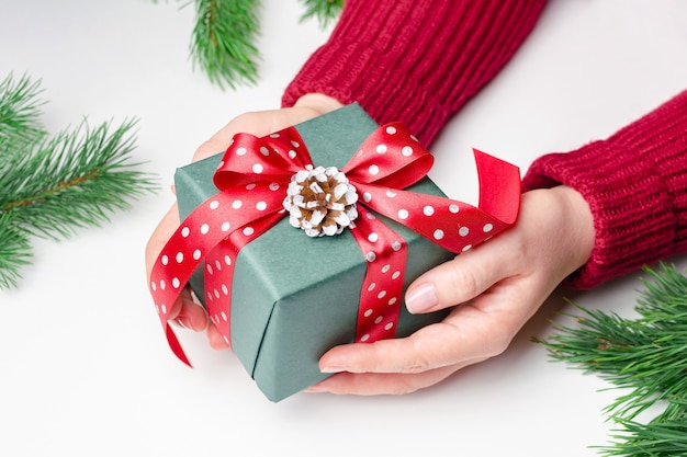 Manos femeninas sosteniendo una caja de regalo de navidad decorada con lazo rojo sobre fondo blanco.