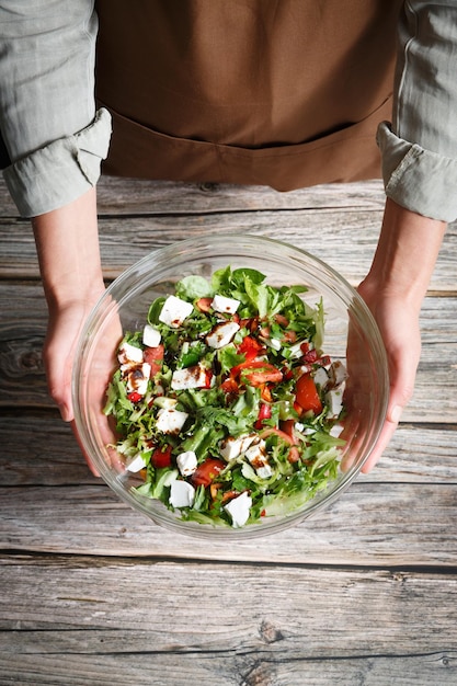 Manos femeninas sosteniendo un cuenco de vidrio de ensalada de verduras con feta sobre un fondo de madera