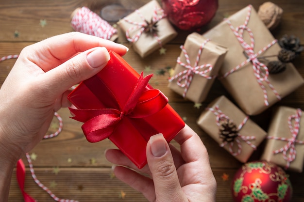 Manos femeninas sosteniendo, envolviendo la caja de regalo de Navidad. Grupo de cajas de regalo envueltas en papel artesanal, adornos rojos, brillo sobre mesas de madera. Chritsmas fondo plano laico.