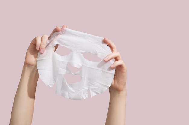 Manos femeninas sosteniendo una hoja de máscara blanca sobre fondo rosa