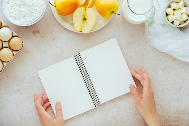 Manos femeninas sosteniendo un libro de cocina abierto con ingredientes harina leche huevos manzanas mantequilla