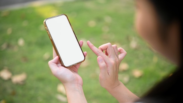 Manos femeninas sosteniendo una maqueta de pantalla blanca de teléfono inteligente contra el primer plano de fondo verde borroso del parque