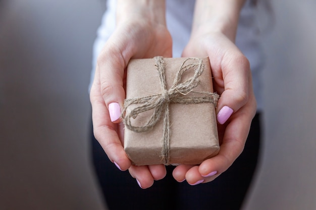 Manos femeninas sosteniendo una pequeña caja de regalo envuelta en papel artesanal con cinta. Pequeño regalo de navidad o año nuevo decorado en manos de mujer. Celebración de cumpleaños de San Valentín de Navidad presente concepto romántico